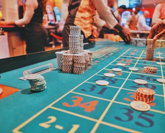 A casino scene with a roulette table features prominently, covered in green felt with colorful numbers and betting squares. Several stacks of poker chips are placed on different numbers, and the hands of several people are visible as they interact with the table. In the background, there are slot machines and more people, creating a vibrant and busy atmosphere.