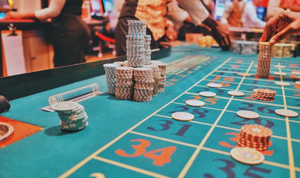 A casino scene with a roulette table features prominently, covered in green felt with colorful numbers and betting squares. Several stacks of poker chips are placed on different numbers, and the hands of several people are visible as they interact with the table. In the background, there are slot machines and more people, creating a vibrant and busy atmosphere.
