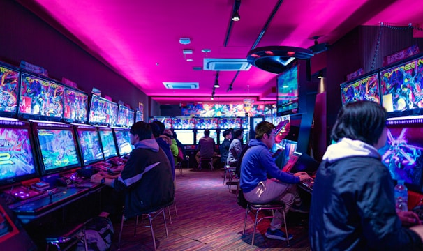 A vibrant arcade with rows of gaming machines adorned with colorful screens. Several people are immersed in playing games, seated on stools in a dimly lit room accented by an intense pink ceiling light. The atmosphere is lively and engaging.