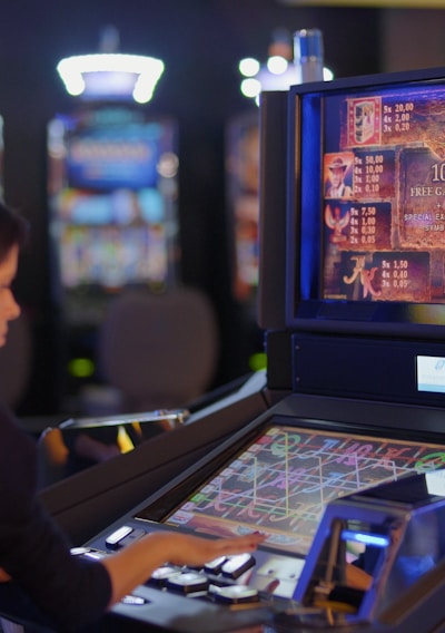 A person is seated at a slot machine console in a casino, focusing intently on the game displayed on the screen. The background shows several other illuminated gaming machines, adding a vibrant and colorful atmosphere to the scene.