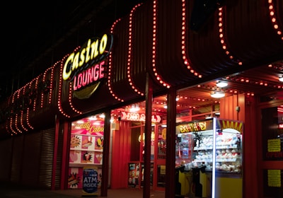 A brightly lit casino lounge entrance adorned with neon lights. The exterior features colorful signage including the words 'Casino' and 'Lounge' in vivid colors, with additional lights framing the building. Inside, a glimpse of a claw machine filled with plush toys and arcade games is visible. The area exudes an inviting yet bustling atmosphere typical of entertainment venues.