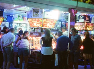 A group of people standing in front of an illuminated amusement arcade, engaging with various gaming machines. The environment is bustling with activity, and colorful lights create a vibrant atmosphere. The ceiling is decorated with large, playful images of dice and other game-related graphics, enhancing the lively mood.